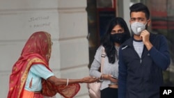 An elderly Indian woman seeks alms as youth wearing pollution masks walk through a shopping area in New Delhi, India, Nov. 14, 2019.