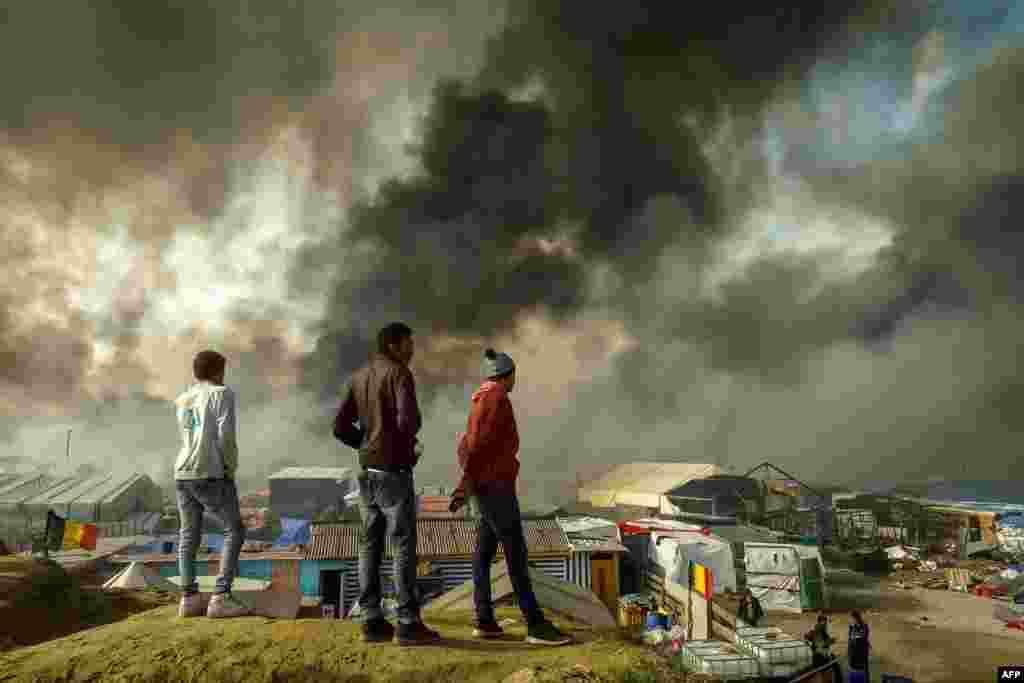 Migrants stand on a hill overlooking the "Jungle" migrant camp in Calais, northern France, as smoke rises during a massive operation to clear the squalid settlement where 6,000-8,000 people have been living in dire conditions.