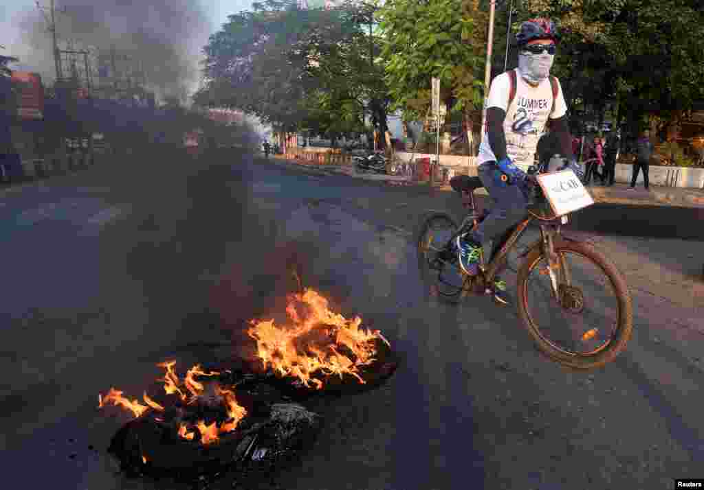 آل آسام چٹیا اسٹوڈنٹس یونین کی جانب سے ترمیمی بل کے خلاف ہڑتال کے دوران ایک سائیکل سوار شخص جلتے ہوئے ٹائروں کے قریب سے گزر رہا ہے۔ سائیکل پر لگے پلے کارڈ پر بھی بل مخالف نعرہ درج ہے۔